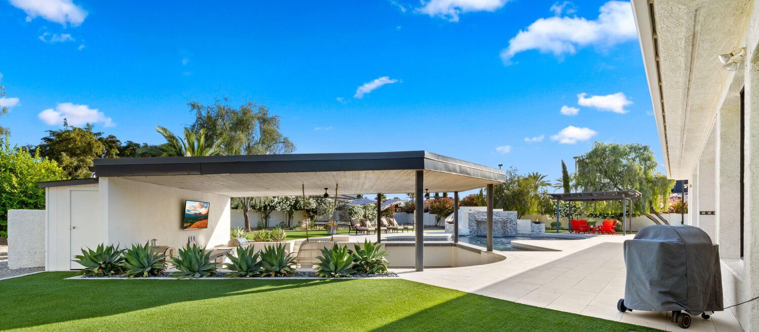 Modern backyard in Glendale, AZ, with a covered patio featuring a TV, surrounded by lush greenery and agave plants. Artificial turf installations enhance the sleek swimming pool and lounge area under a pergola. The clear blue sky enhances the tranquil setting.