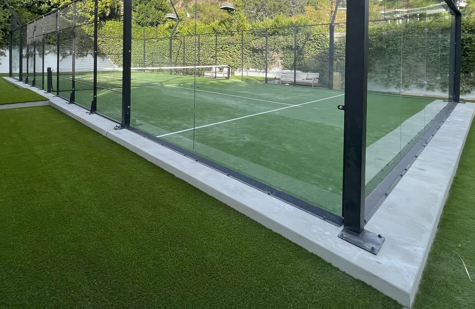 A padel court in Glendale, AZ, surrounded by glass walls and featuring a green synthetic turf with white boundary lines, is set outdoors against a backdrop of lush greenery.