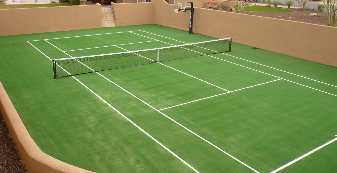 Outdoor court in Glendale AZ featuring tennis and basketball setups on vibrant artificial sports turf, enclosed by tan walls with white boundary lines. A basketball hoop stands at one end, while a tennis net stretches across the center. Desert plants beautifully adorn the surroundings.