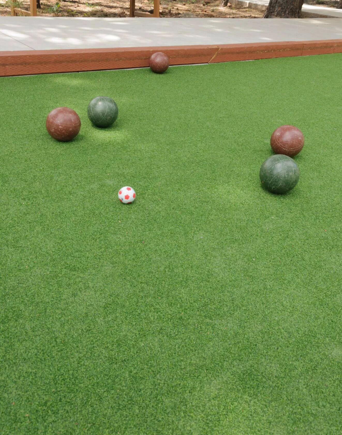 A grass bocce court with four large bocce balls in red and green scattered across the synthetic turf. A small white ball with red dots is positioned in the center. The background features a walkway and a tree, showcasing a perfect installation in Glendale, AZ.