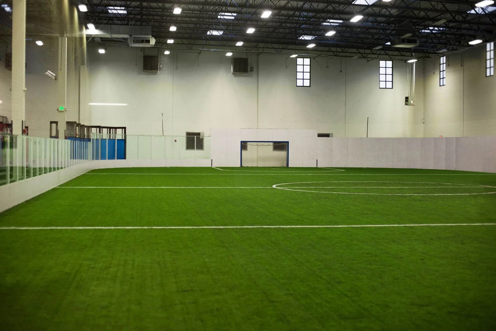 Indoor soccer field with artificial sports turf, white walls, and a high ceiling with industrial lights. The field has clear glass barriers along the sides and a goal at the far end. Several windows let natural light in. Located conveniently in Glendale, AZ, it's a premier spot for enthusiasts.