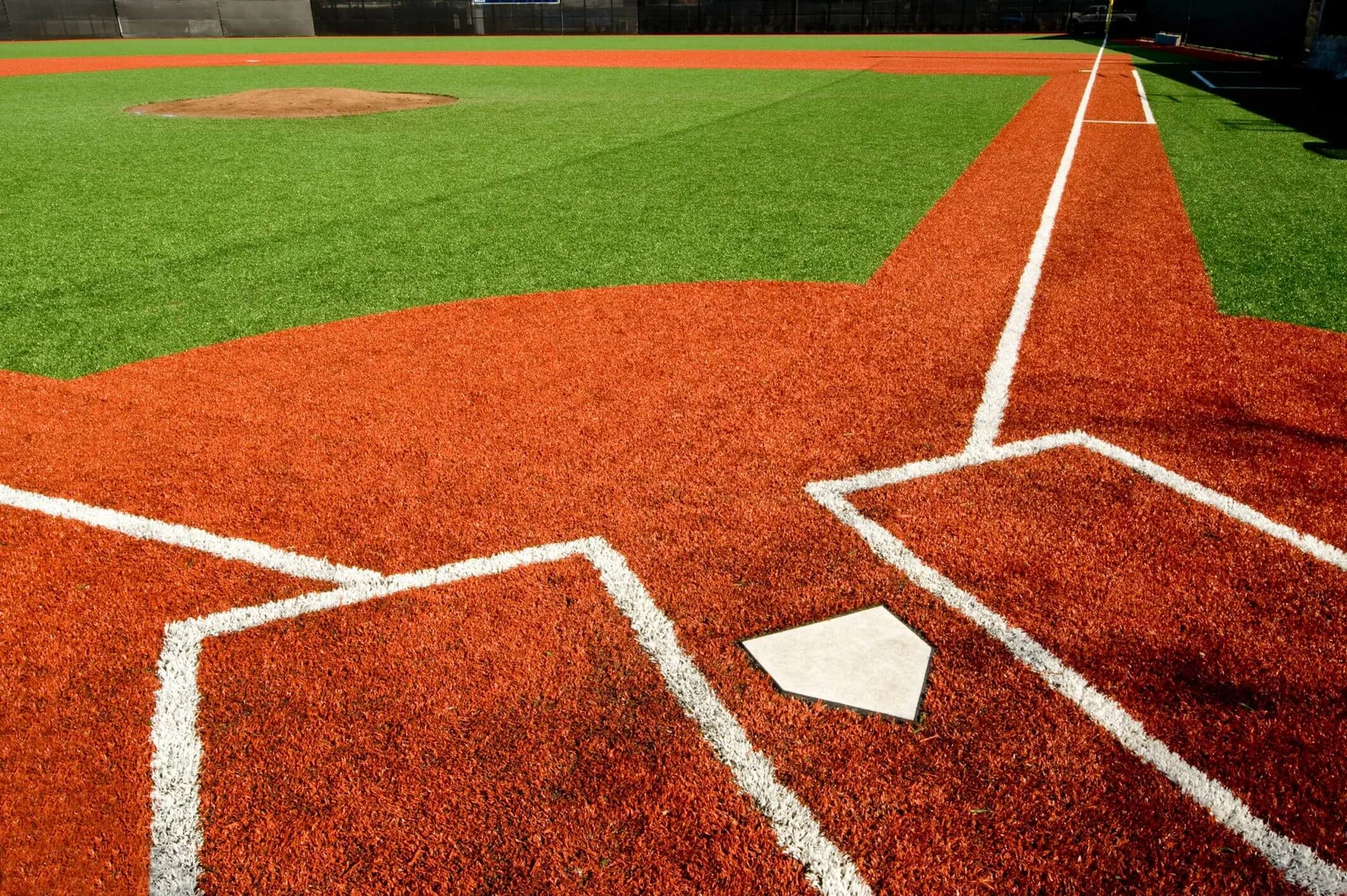 A close-up view of a baseball field in Glendale, AZ shows the home plate and surrounding dirt area in the foreground. Red synthetic turf covers the base paths, contrasted with green artificial sports turf in the field beyond the pitcher's mound.