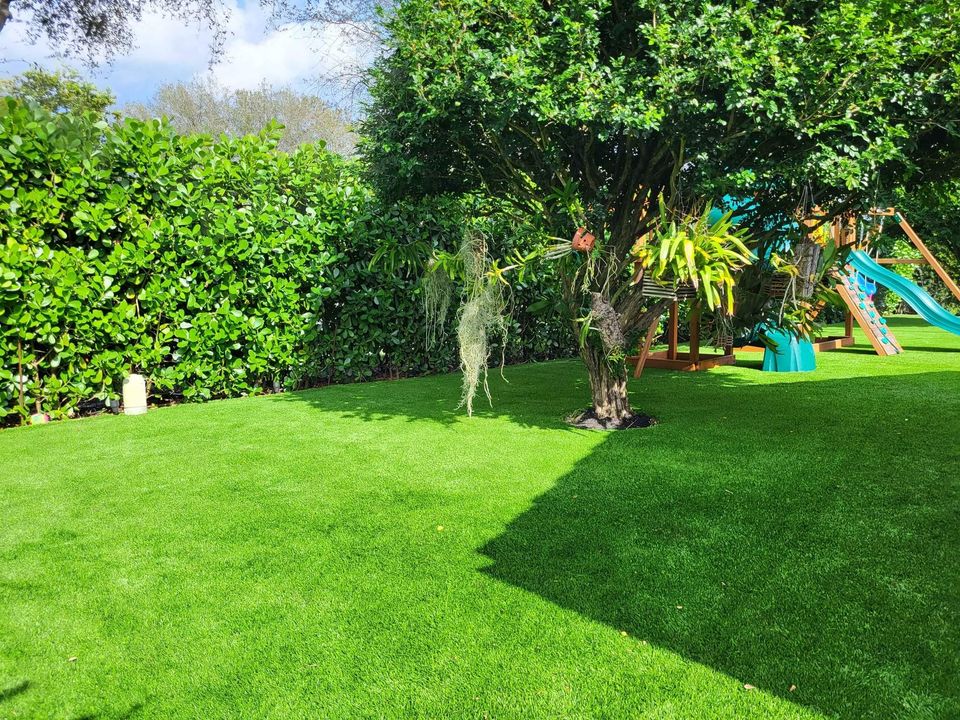 A lush green backyard in Glendale, AZ, features pet-friendly artificial turf and a playset, surrounded by tall hedges. Bright sunlight casts shadows on the well-maintained grass. A slide is partially visible in the background, and the sky is partly cloudy.