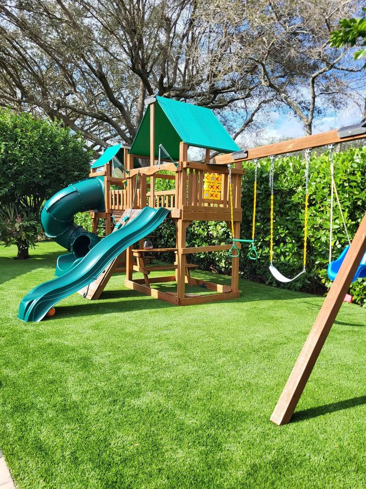 A wooden playground set with a green slide, spiral tube slide, swings, and climbing area is nestled on vibrant artificial turf installations. Tall trees and bushes surround the area under a clear blue sky in Glendale, AZ.