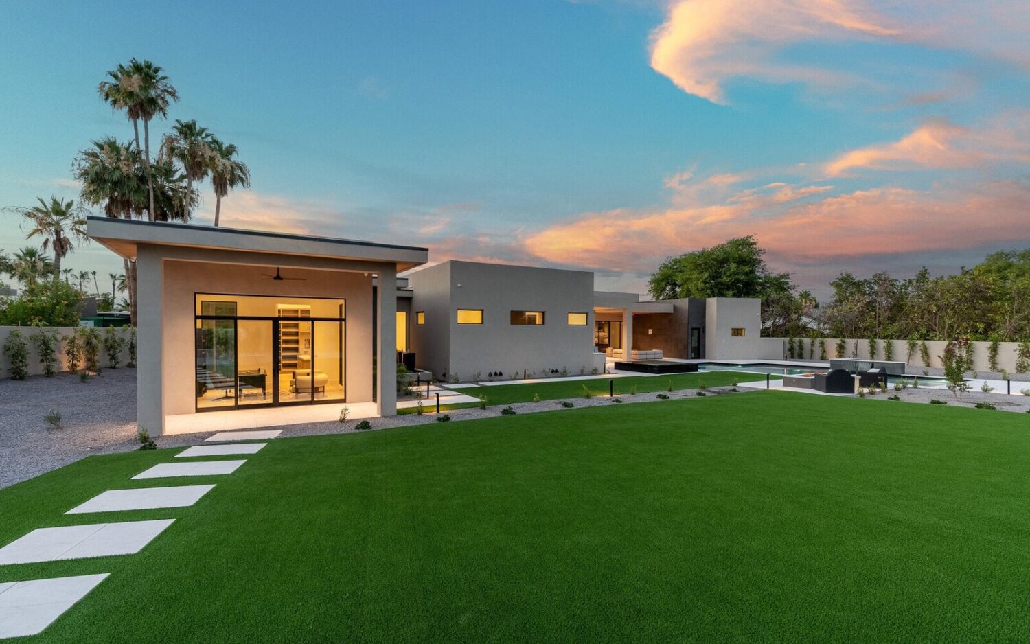 Modern single-story house with expansive glass windows, surrounded by a meticulously kept lawn of artificial turf and stepping stones. Tall palm trees stand in the background under a blue sky with pink clouds during sunset, making this Glendale, AZ gem truly captivating.