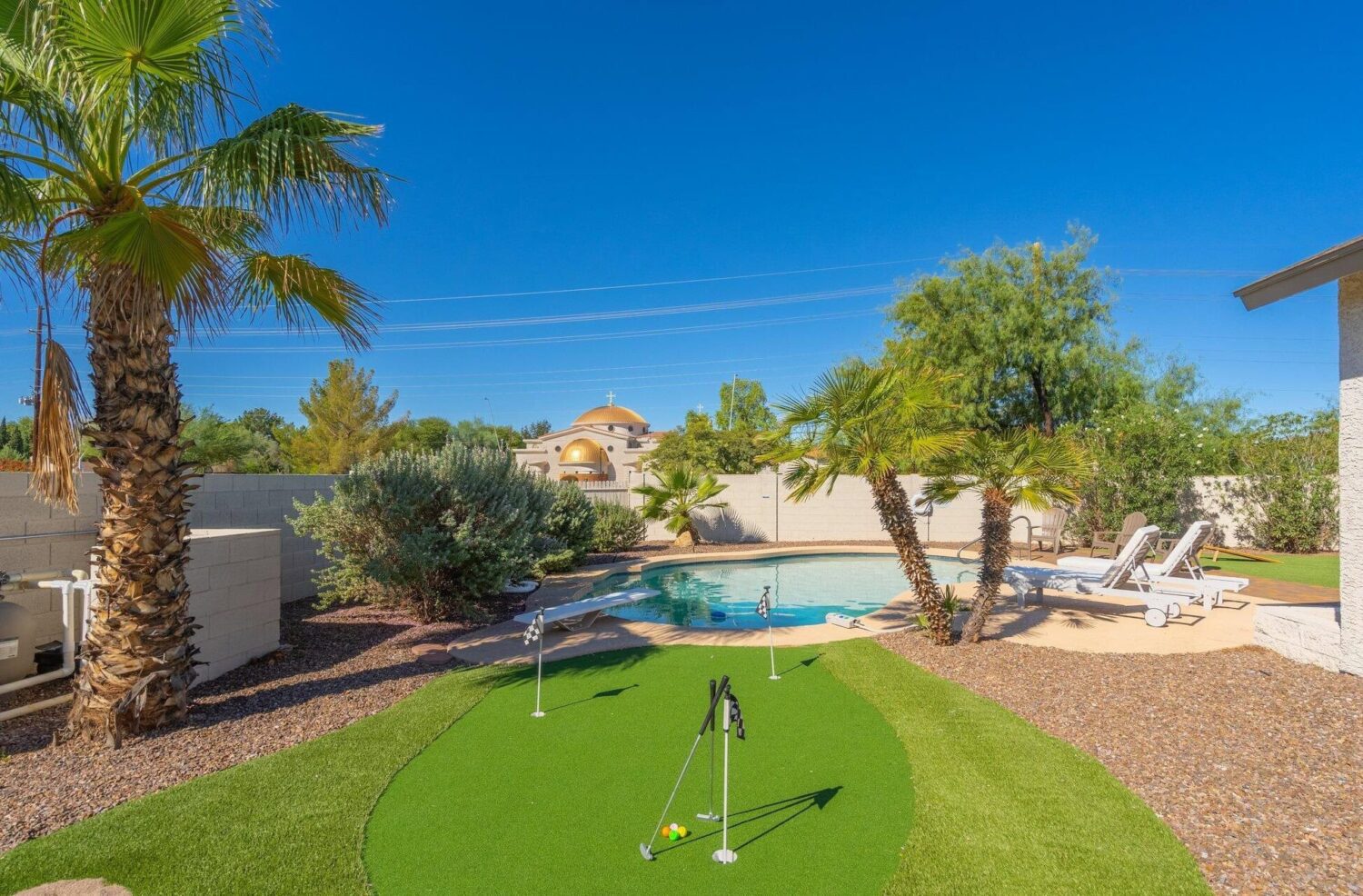 A backyard oasis in Glendale, AZ, featuring a small circular swimming pool surrounded by palm trees and sun loungers. In the foreground, you'll find synthetic putting greens with flags made from premium artificial turf. The scene is set under a clear blue sky.