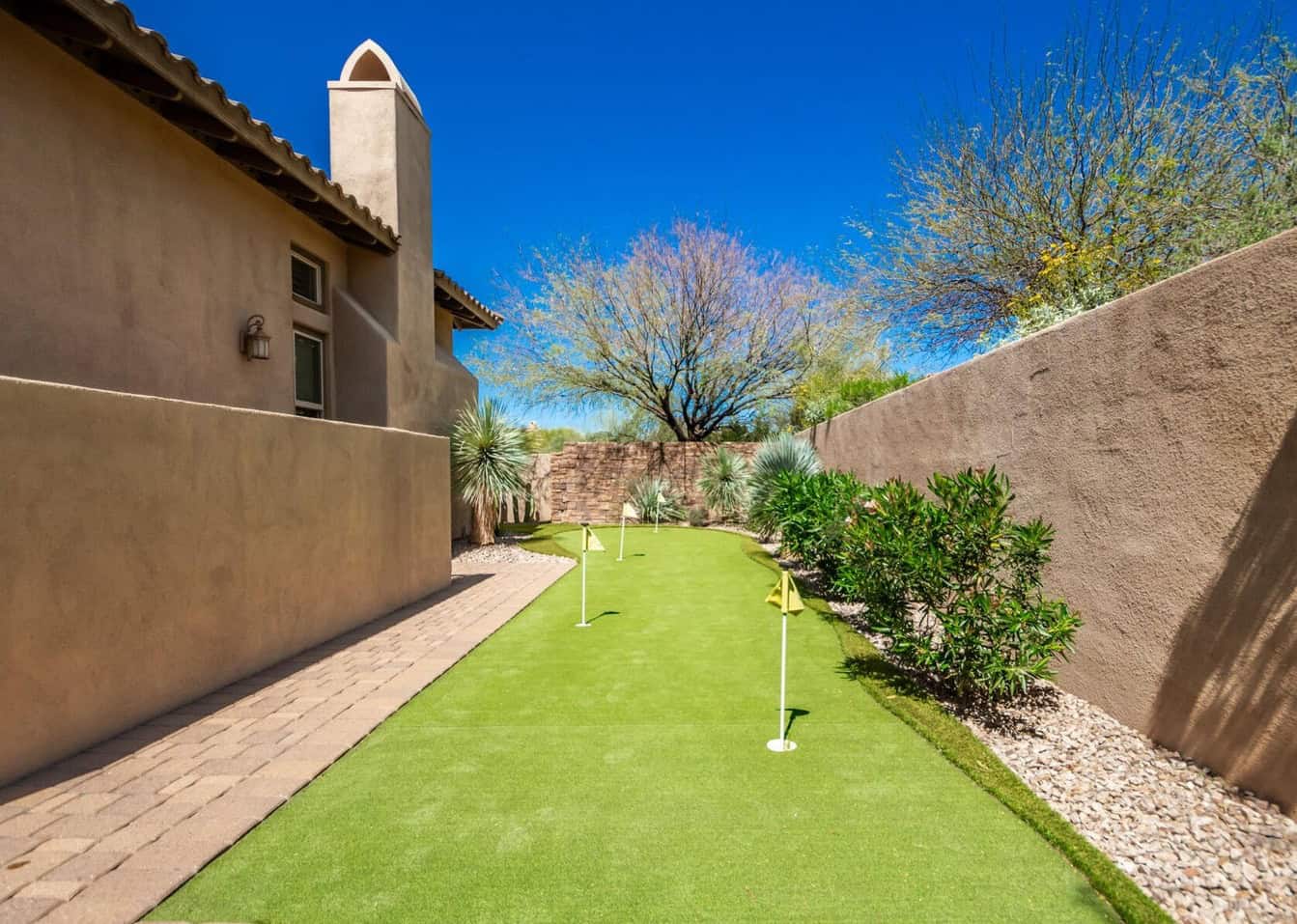 A narrow backyard features a well-maintained synthetic putting green with three holes, bordered by a stone walkway and walls. The artificial turf complements the desert plants and trees, all visible under Glendale AZ's clear blue sky.