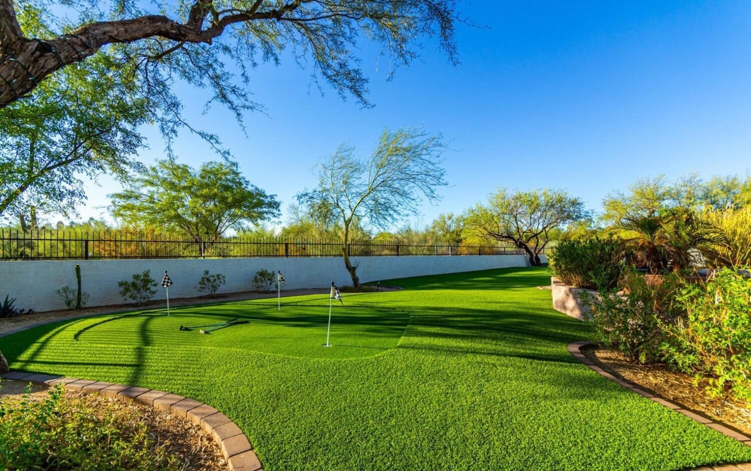 In a sunny Glendale, AZ backyard, a small synthetic putting green crafted from artificial turf boasts several holes, surrounded by trees and a white fence. The yard's blue sky backdrop complements the landscaped plants along the perimeter.