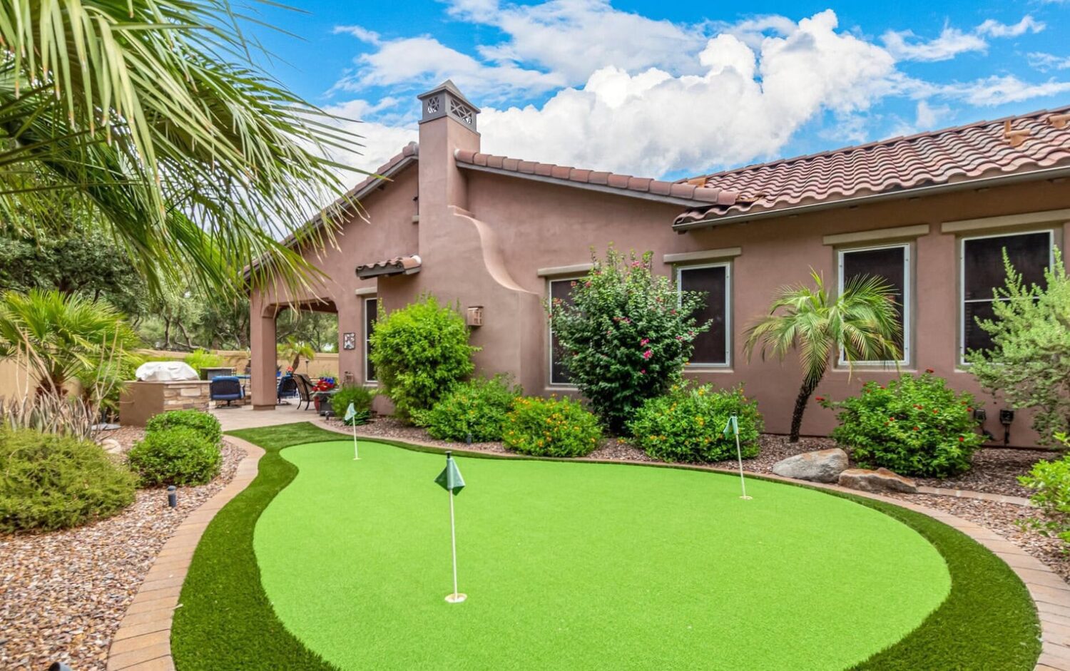 A backyard scene in Glendale, AZ showcases synthetic putting greens surrounded by desert landscaping and small palm trees. The house in the background boasts stucco walls and a red-tiled roof beneath a bright blue sky with fluffy clouds, complementing the vibrant artificial turf.