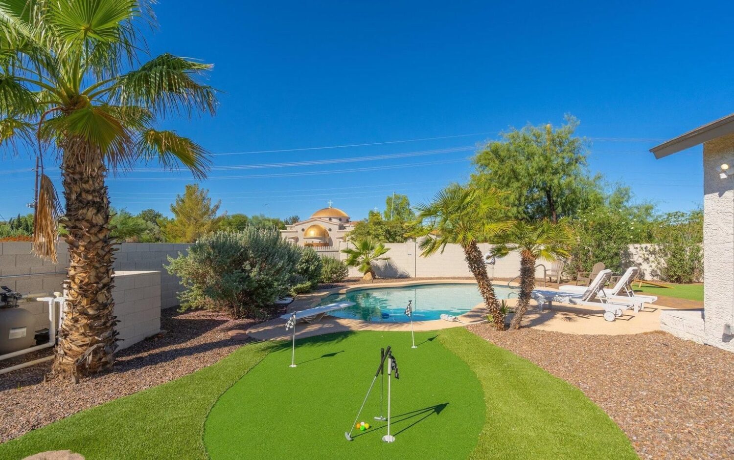 A backyard oasis in Glendale, AZ, featuring a small artificial putting green installed by expert artificial grass installers. Palm trees sway beside a shimmering swimming pool with sun loungers. Enclosed by a stone wall, this serene escape sits under a clear blue sky.